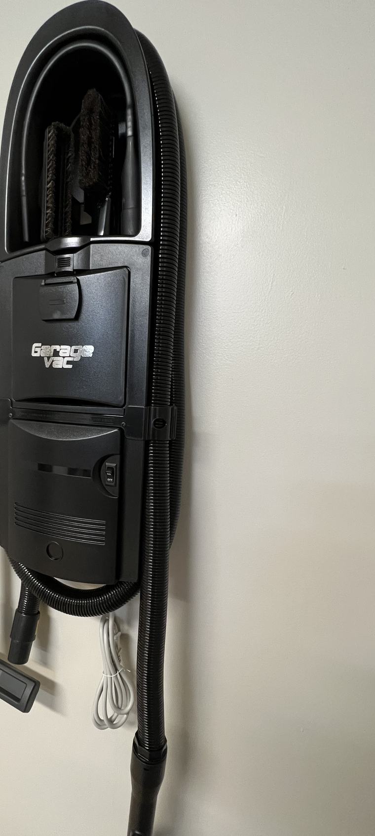 A GarageVac in black mounted on a garage wall, hose wrapped around the body and tools stored in the caddy at the top.